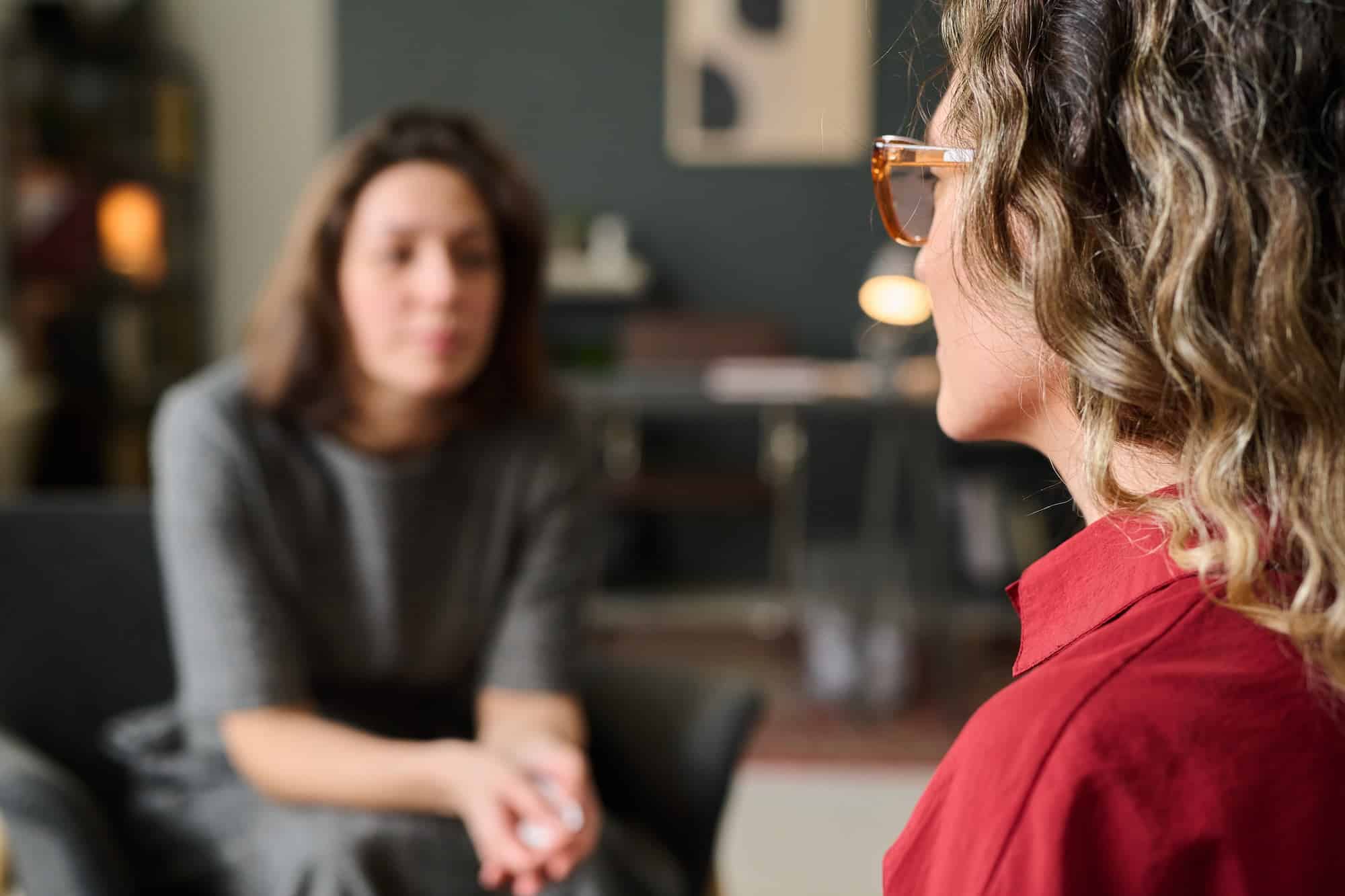 A therapy session where two women are engaged in conversation. The woman in the foreground, wearing glasses and a red blouse, listens attentively while the other woman, slightly blurred, appears to be speaking in a supportive environment.