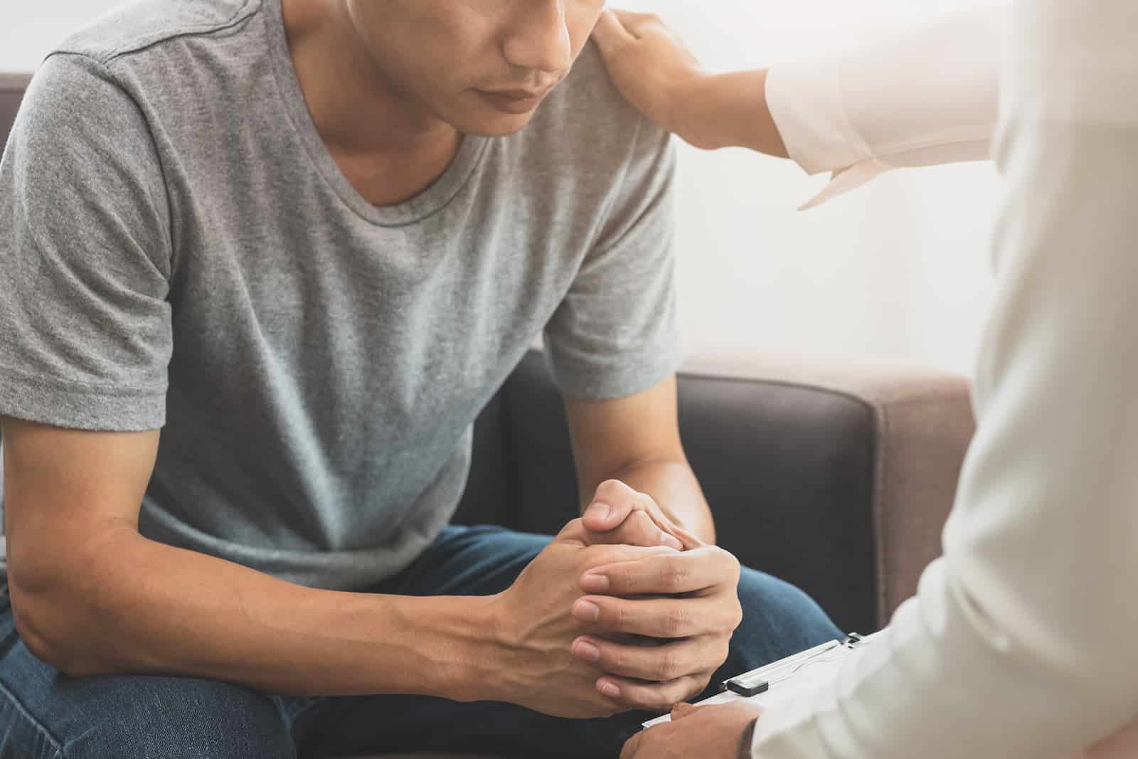 A man wearing a gray t-shirt sits on a couch with his hands clasped together, looking down in contemplation. A therapist or support person, partially visible in the foreground, places a reassuring hand on his shoulder, offering comfort and support during the session.