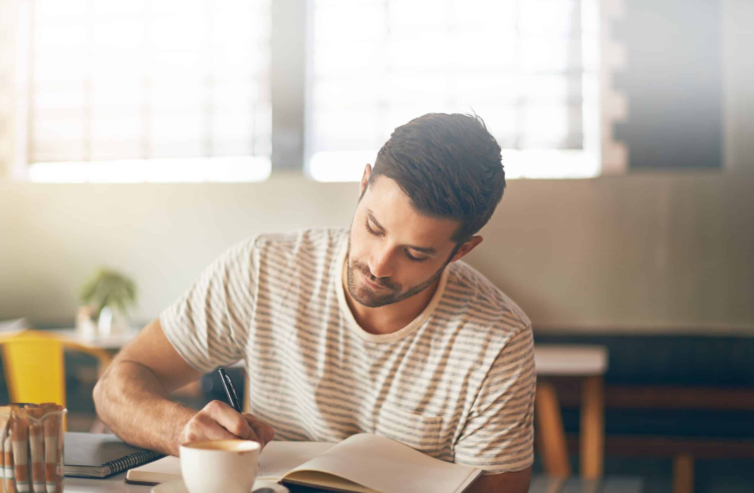 A man in a well-lit coffee shop journalling about his anger. He writes in pen and has a hot coffee next to him.