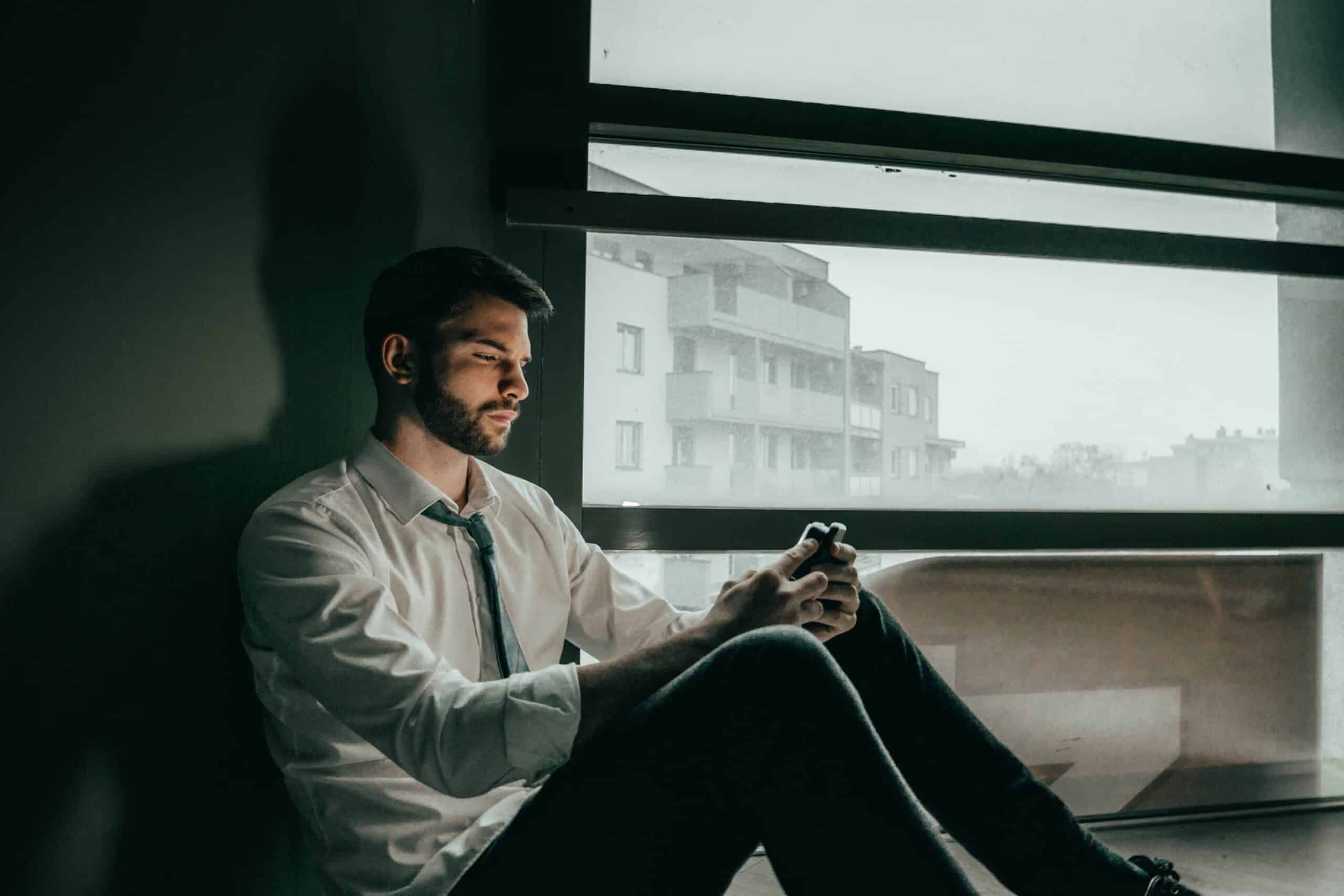 A caucasian man sitting on the floor inside a dark room. His face is lit by the light of a cellphone he stares at. A window shows a foggy day outside.